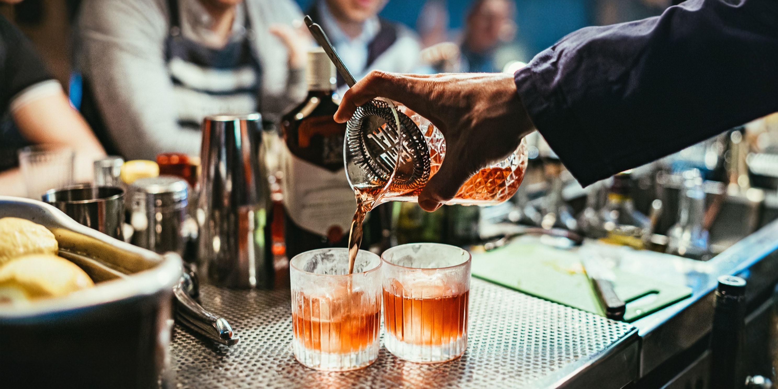 Bartender straining a stirred cocktail into two rocks glasses at a bar